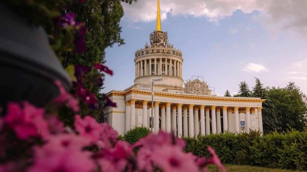 Квест на ВДНГ: 20+ локацій і подарунки за печатки Головний павільйон ВДНГ зі шпилем на тлі неба, у розмитому передньому плані рожеві квіти петунії