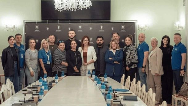 КМДА відкрила центр для літніх людей, людей з інвалідністю Група людей стоїть у залі для нарад перед довгим столом на фоні банера Київської міської адміністрації