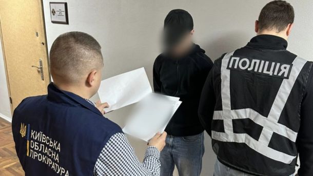 Викрали військового та вимагали 30 тисяч: групу затримали на Київщині Працівник прокуратури зачитує щось з листків формату А4 підозрюваному, який стоїть в приміщенні біля стіни, біля підозрюваного також стоїть працівник поліції