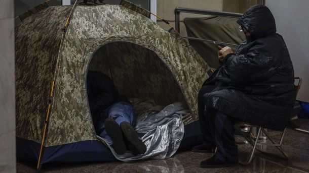 Знову погодинні відключення світла в Києві: стабілізують до 22:00 Намет на станції метро, там спить чоловік, поряд з наметом стоїть стілець і на ньому сидить жінка в теплому одязі