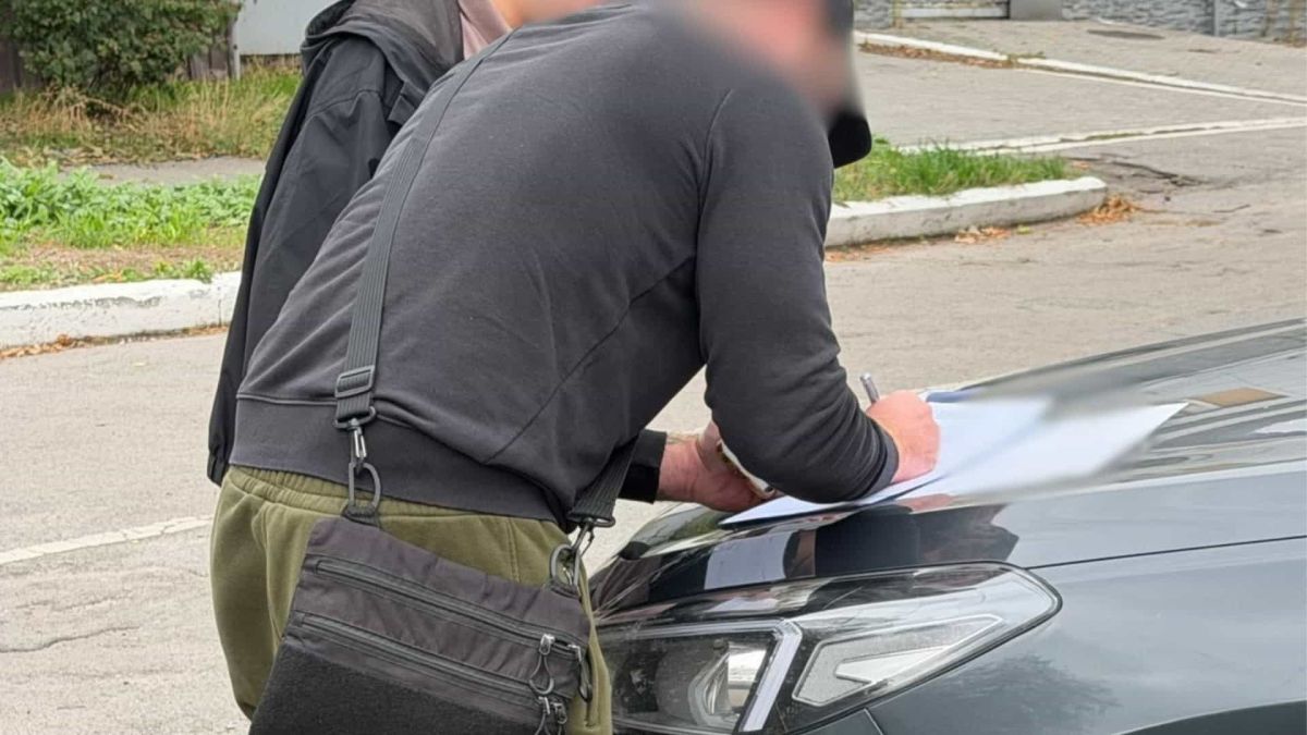 Модульний будинок за 12 тис. доларів не встановили: директора судитимуть Чоловік із заблюреним обличчям підписує документи на капоті автомобіля у справі про невстановлений модульний будинок у Києві