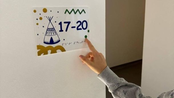 У Києві змінять маршрути 9 автобусів через ярмарки 18–19 жовтня Дитяча рука, біля стіни з картинкою, на якій шрифтом брайля написаний зміст картинки
