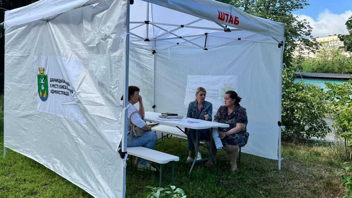 У Києві відкрили штаби допомоги для постраждалих від атаки РФ: адреси та контакти Троє жінок сидять за столом під білим наметом із написом «ШТАБ»