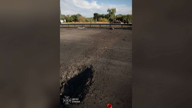 Атака на Київщину: пошкодження у трьох районах Обгоріла ділянка асфальту на проїзній частині, огороджена сигнальною стрічкою, на фоні — люди й дерева