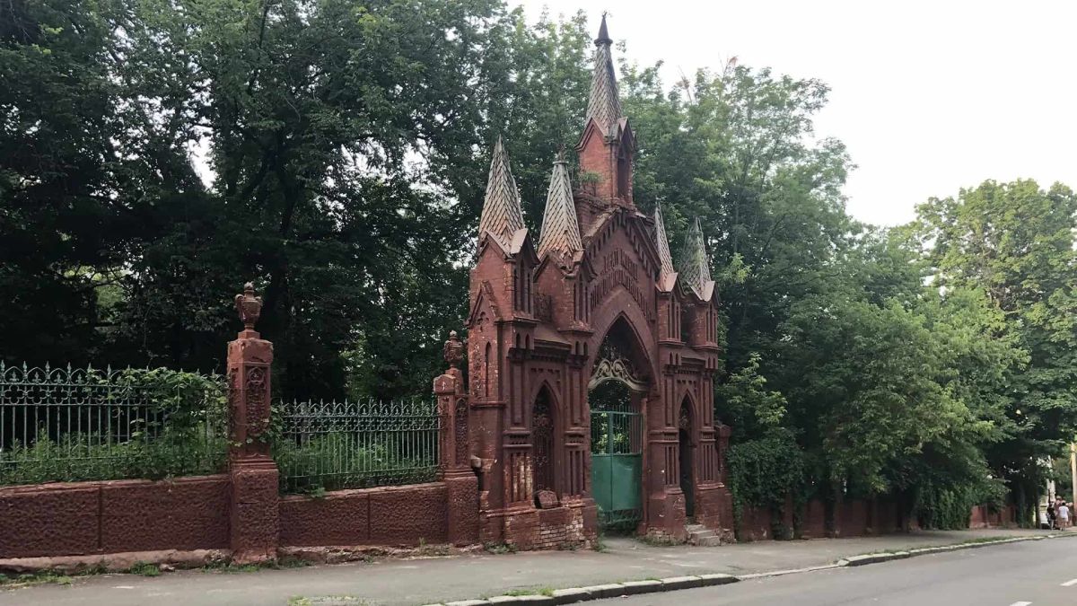 Паркан Байкового кладовища потребує ремонту як підпірна стінка Історична цегляна брама з готичними шпилями біля входу на Байкове кладовище в Києві, зелена огорожа і дерева навколо