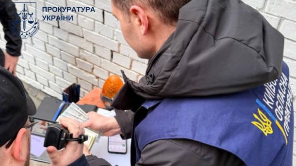 У Києві затримали начальника відділу ДПС за вимагання $500 щомісяця Представник Київської обласної прокуратури у синьому жилеті оглядає вилучені долари США під час затримання начальника відділу ДПС за вимагання хабаря