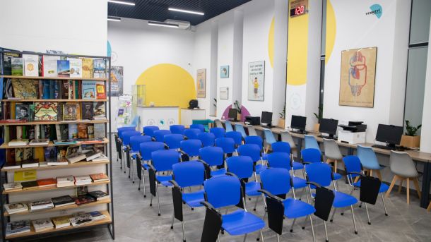 Що купували на «Книжковій країні»: серед лідерів Карпа, Шкляр, Дашвар і Коллін Гувер Фото приміщення з кількома десятками синіх стільків, полицею з книжками та столами з компʼютерами