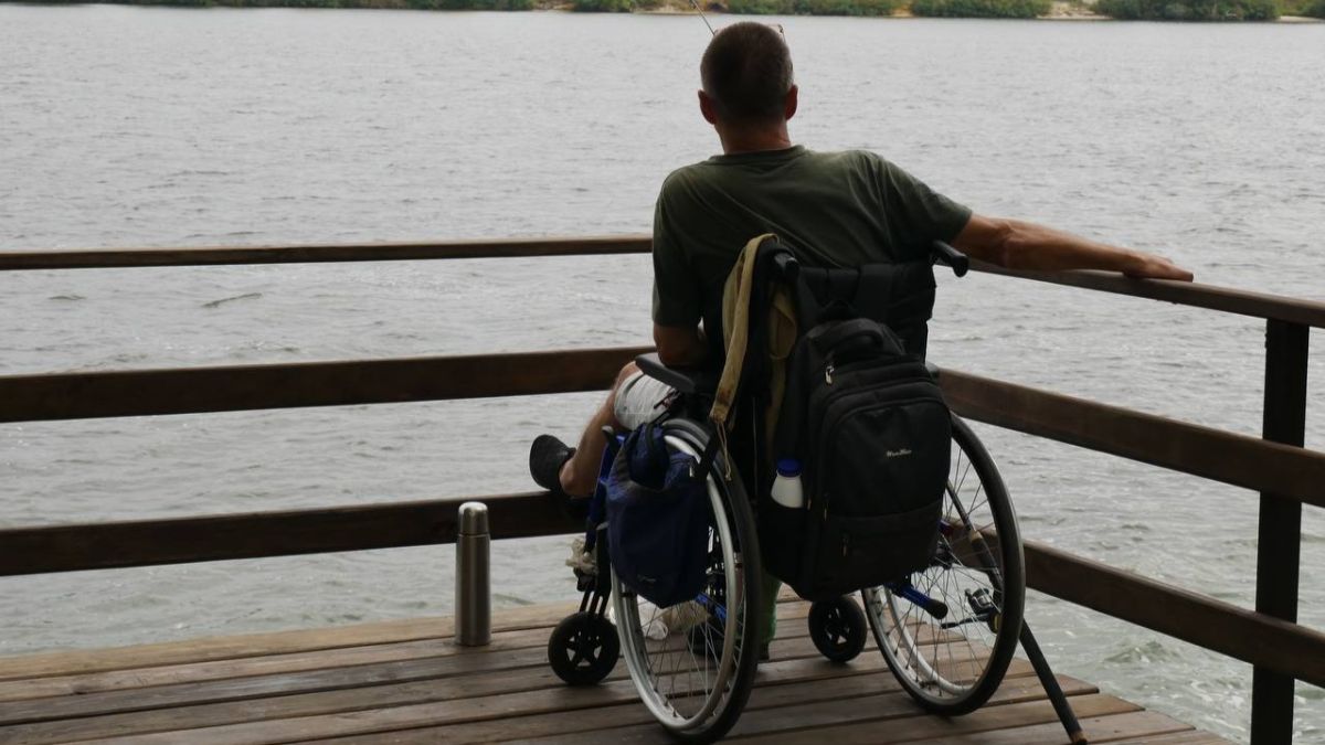 У Києві запустили нові безоплатні програми реабілітації для військових та їхніх родин Чоловік-ветеран з інвалідністю у кріслі колісному сидить на дерев'яному пірсі біля річки