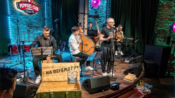 У Херсоні відзначають четверту річницю протестів проти окупантів Музиканти виступають на сцені Peppers Club під час благодійного концерту «НаШапку», на передньому плані — ящик з написом «На перемогу!» для збору донатів