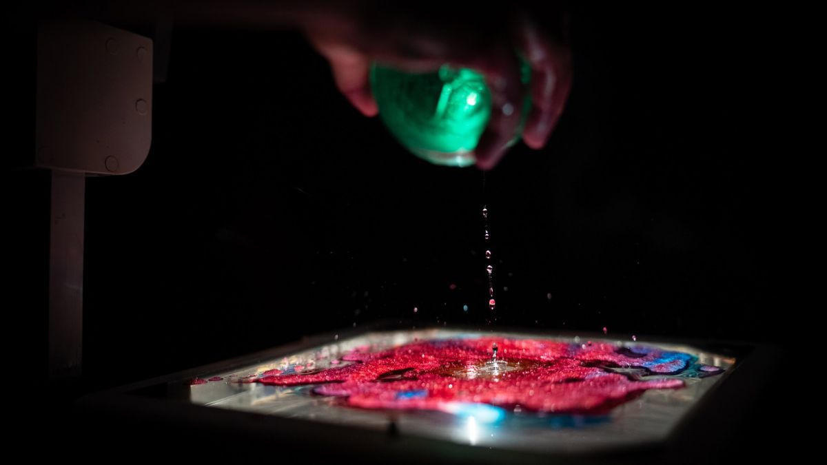 У Києві відкрили виставку художників-військових «Проєкт 24022022» Художня інсталяція на виставці «Проєкт 24022022»: рука художника-військовослужбовця наливає рідину зі світної зеленої посудини на яскраву червоно-синю композицію, підсвічену знизу