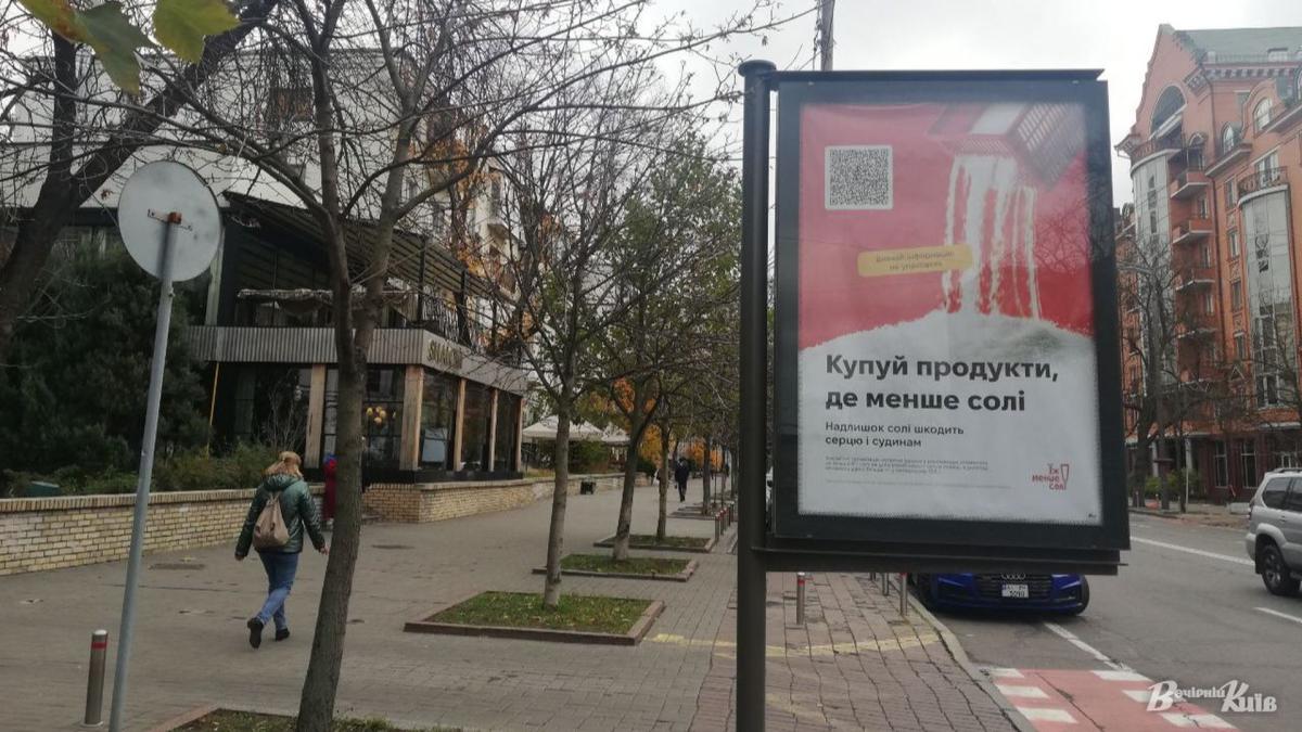 Білборд із закликом "Купуй продукти, де менше солі" на міській вулиці, обабіч якої ростуть дерева