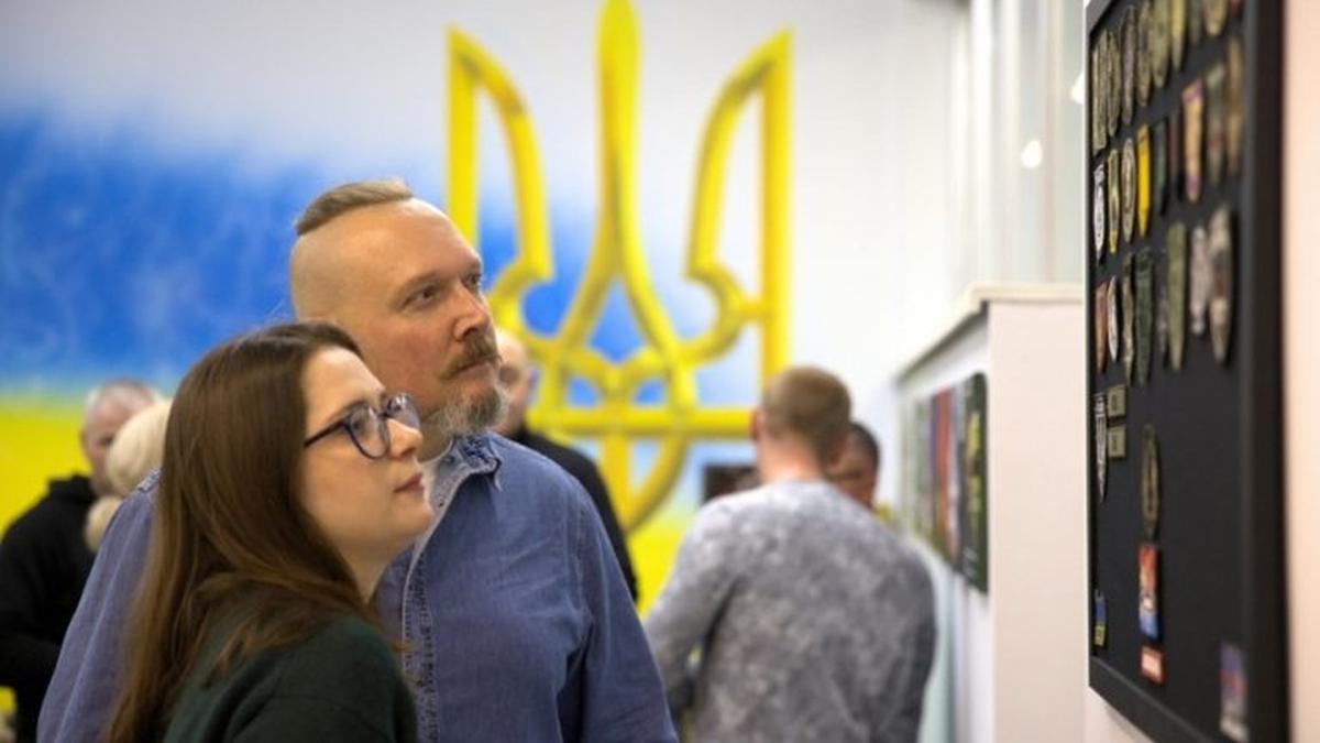 Двоє людей розглядають експонати на стіні; на тлі велике зображення Герба України