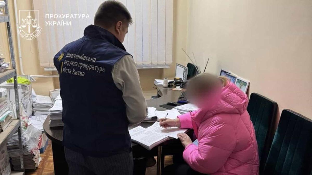 Правоохоронець стоїть біля жінки в рожевій куртці яка за столом підписує папери