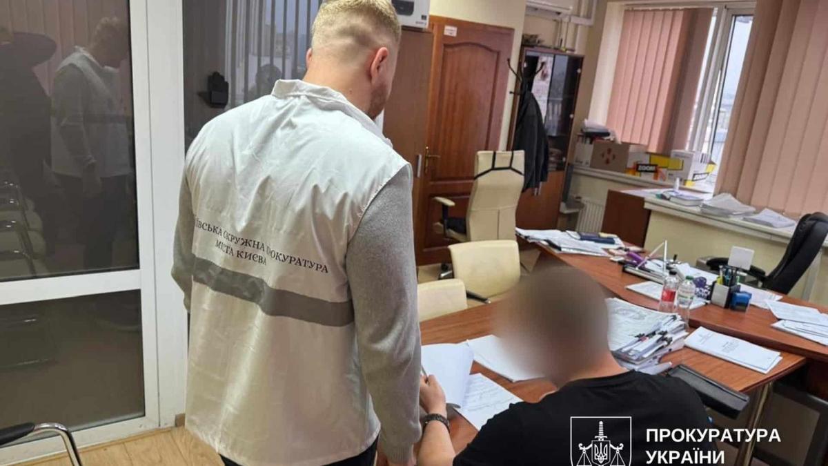 Розкрадання 1 млн грн на будівництві траси ДЮСШ у Києві Працівник прокуратури стоїть біля підозрюваного в кабінеті, де той підписує папери сидячі за столом