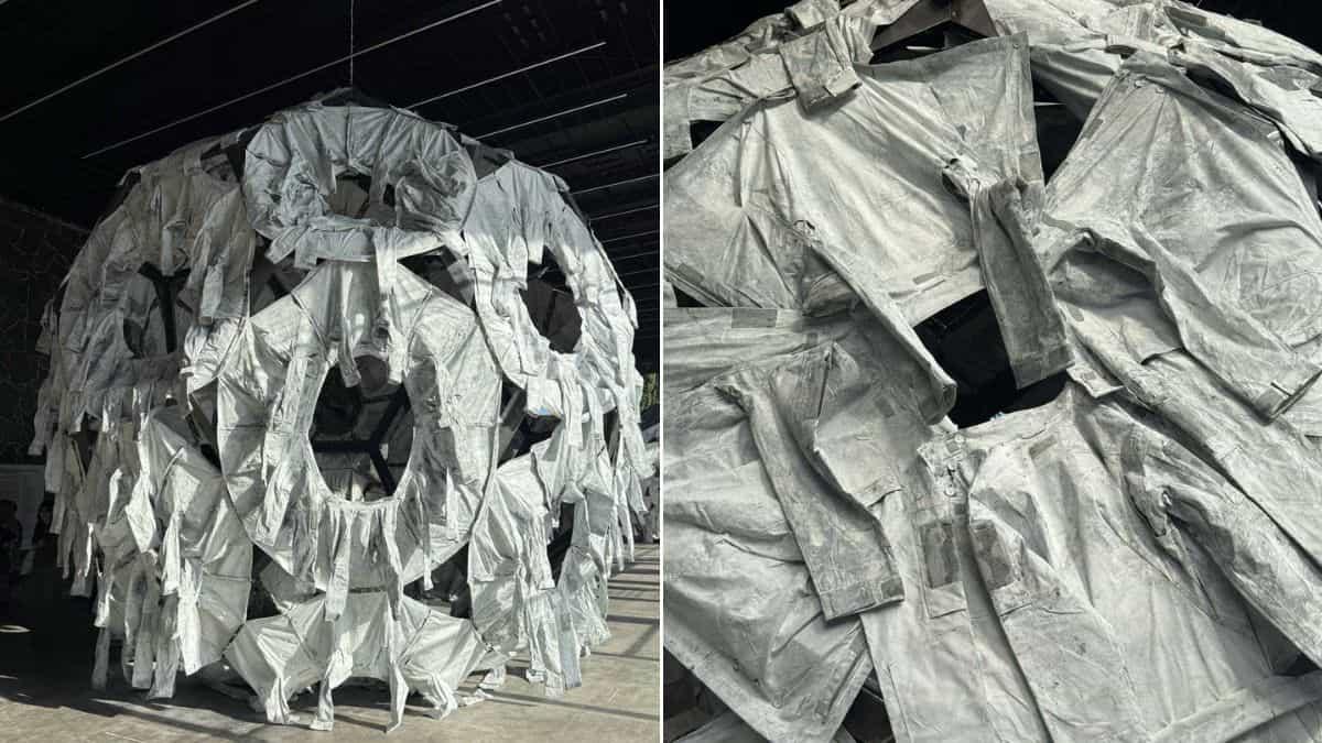 Велика інсталяція у вигляді кулі, складеної з численних сірих захисних костюмів або комбінезонів, підвішена в просторому залі