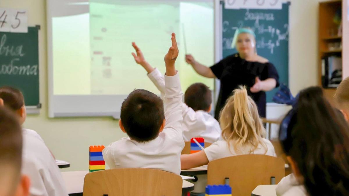 Клас із дітьми, які піднімають руки на уроці, і вчителькою біля дошки з проєктором