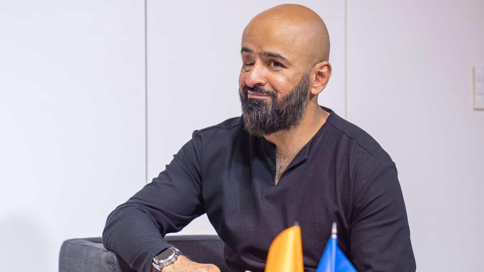 Масі Найем сидить на стільці і посміхається, біля нього український прапор на столі