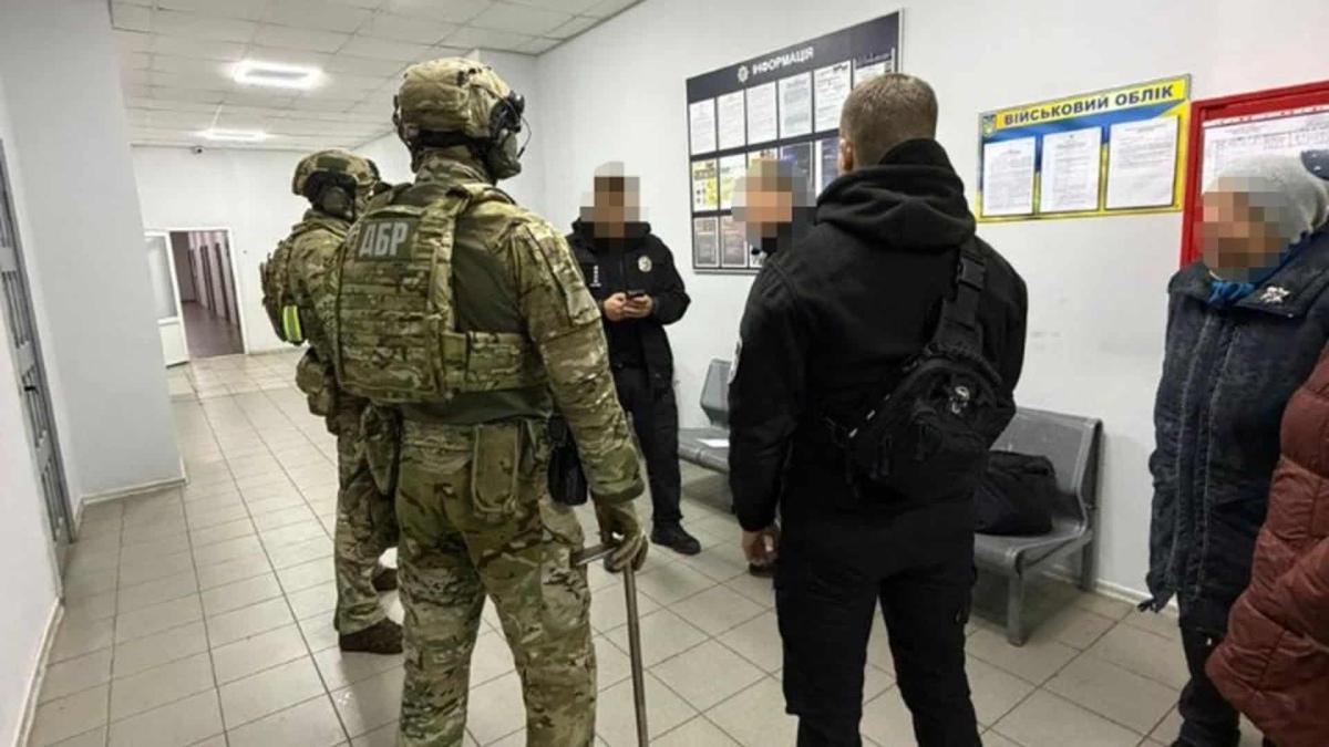 На Київщині поліцейський покривав наркобізнес за $10 тис Працівники ДБР стоять з працівниками поліції в приміщенні, схожому на поліцейський відділок, двоє чоловіків у повному бойовому екіпіруванні