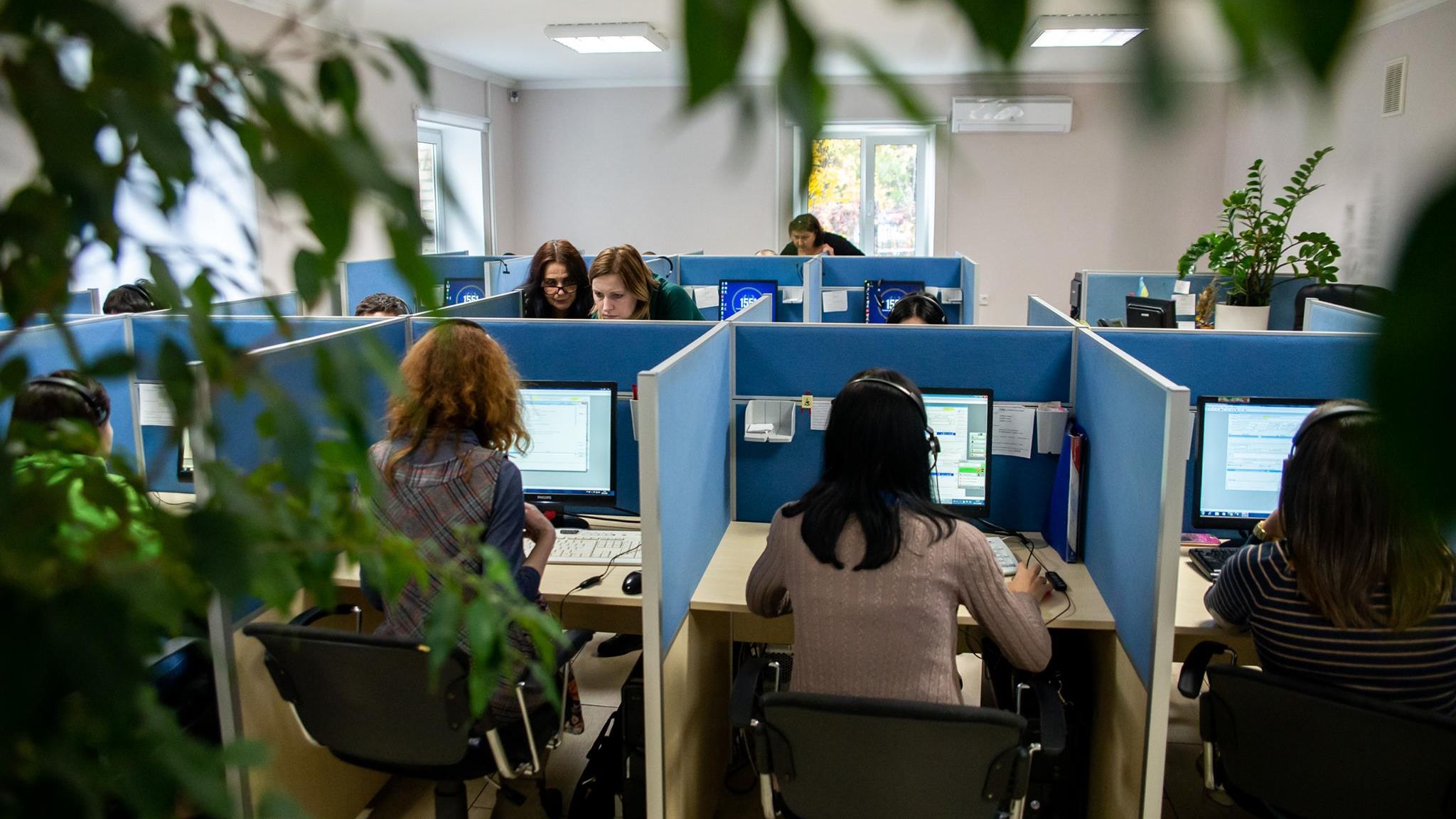 Офісне приміщення з рядами робочих місць, розділених синіми перегородками. Люди сидять за комп’ютерами з гарнітурами, працюючи в кол-центрі.
