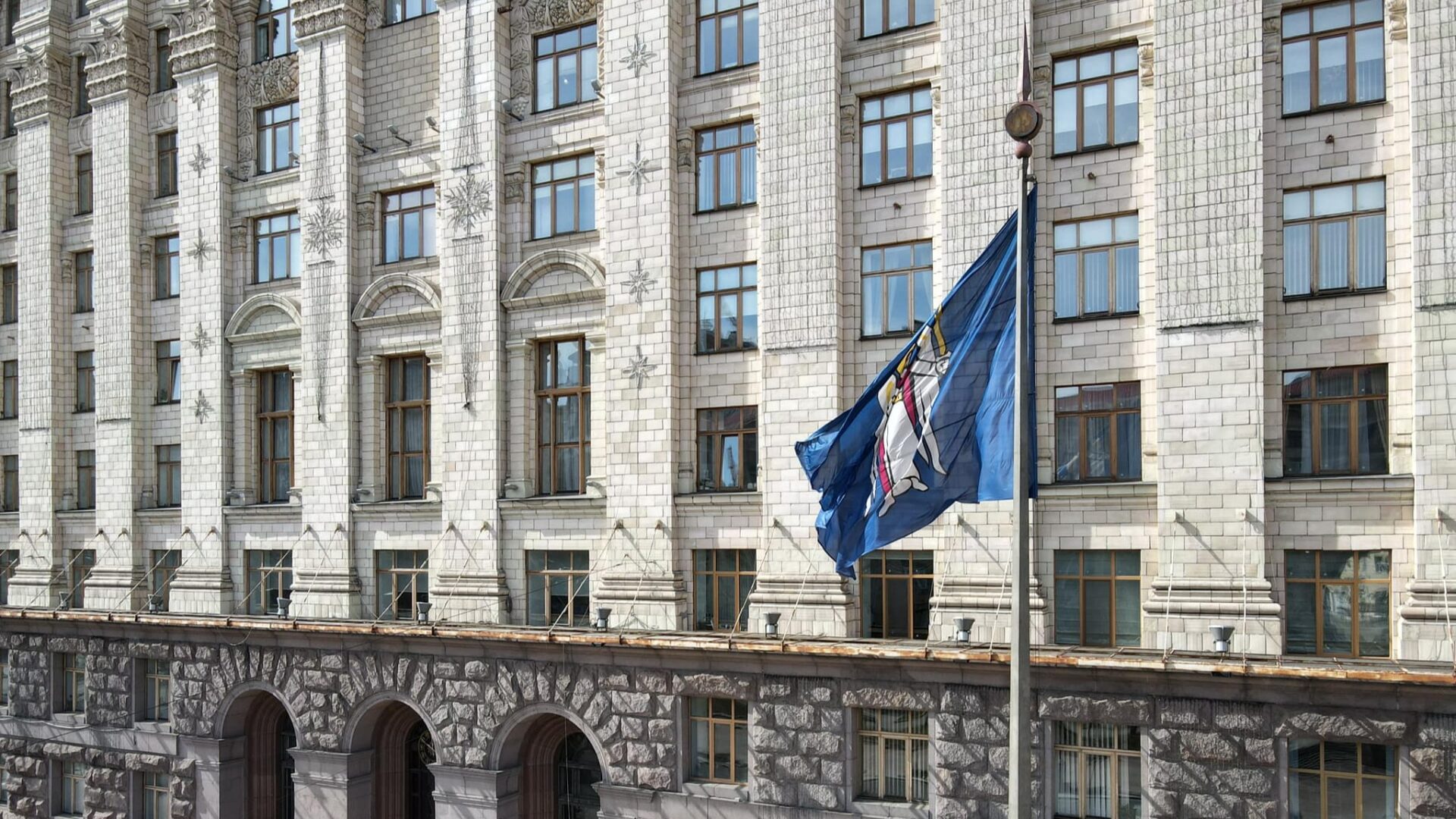 Фасад будівлі КМДА з білої плитки з великими вікнами, перед якою на флагштоці майорить синій прапор із гербом Києва