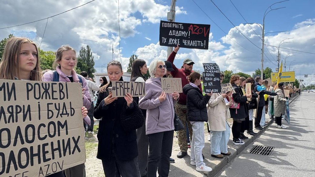 Група людей з плакатами стоїть уздовж дороги під час протестної акції в сонячний день