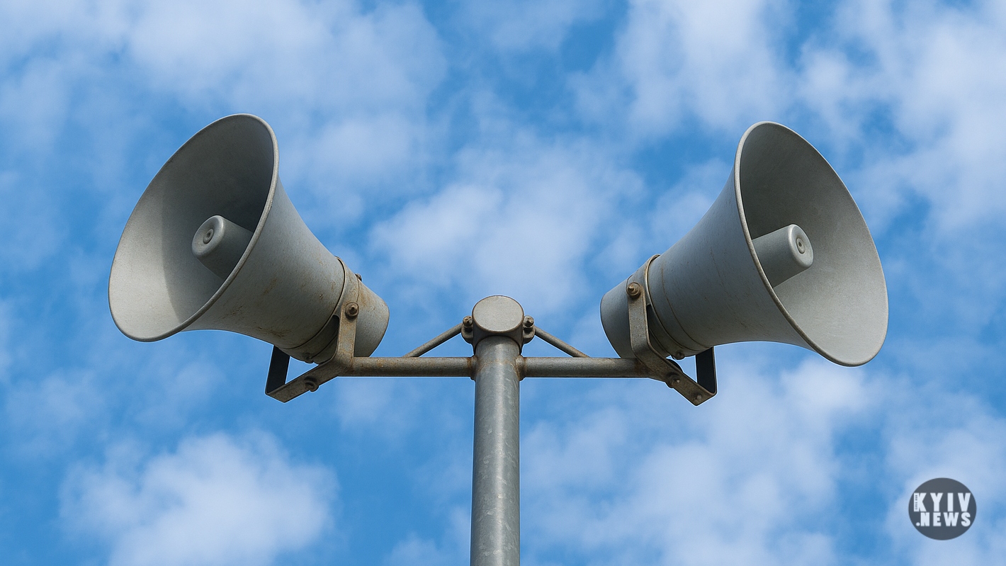 Дві сірі гучномовці на стовпі спрямовані у різні боки на фоні блакитного неба з хмарами