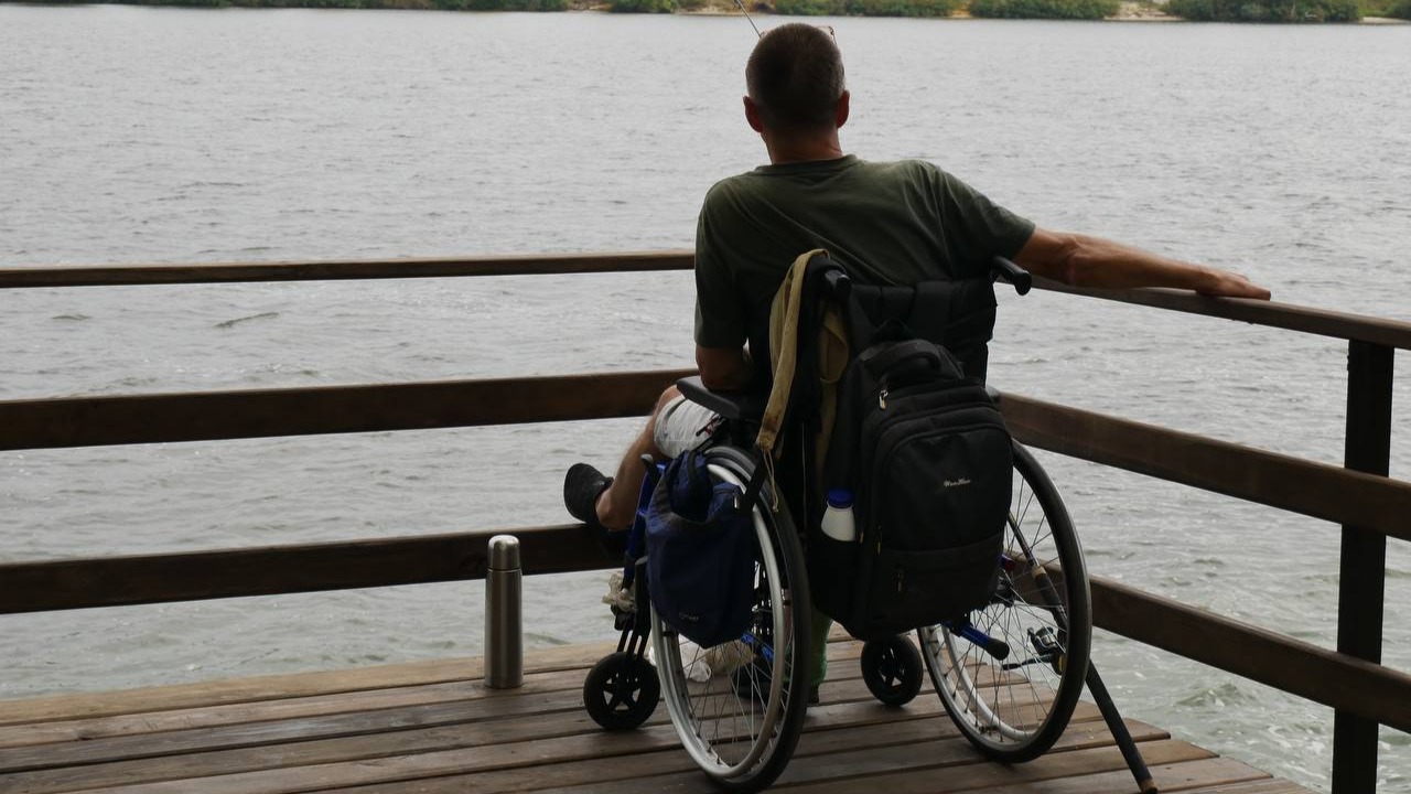 Чоловік-ветеран з інвалідністю у кріслі колісному сидить на дерев'яному пірсі біля річки