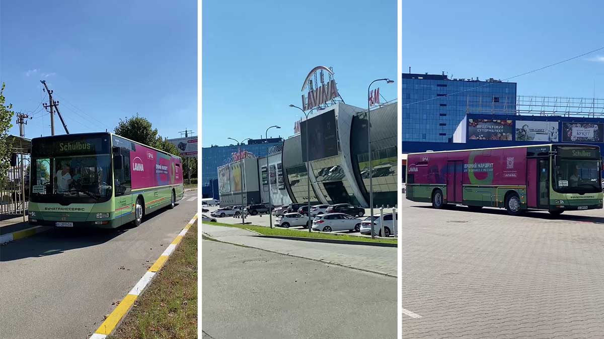 Зелений міський автобус біля зупинки на вулиці Києва, сучасна архітектура торгового центру з логотипом "Київ" на фасаді, автомобілі та житлові будівлі у фоновому плані, ясна погода.