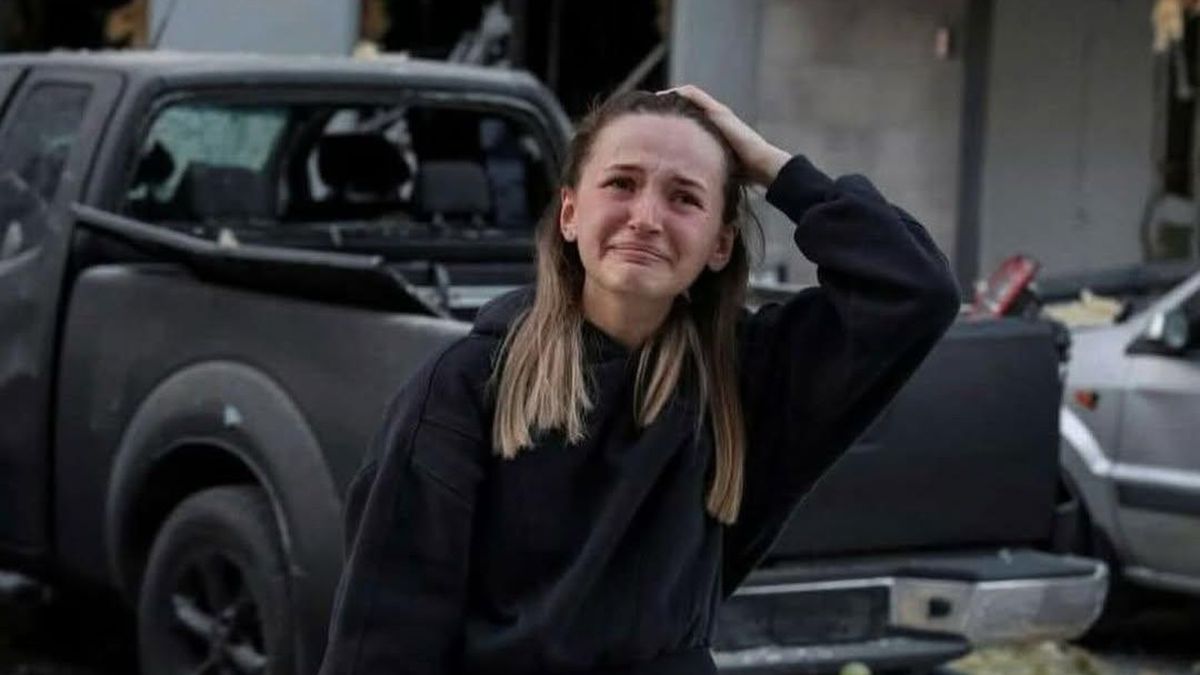 Жінка у жалобній позі, з сумом на обличчі, стоїть на фоні зруйнованих автомобілів біля будівлі, що відображає трагічні наслідки конфлікту в Києві.