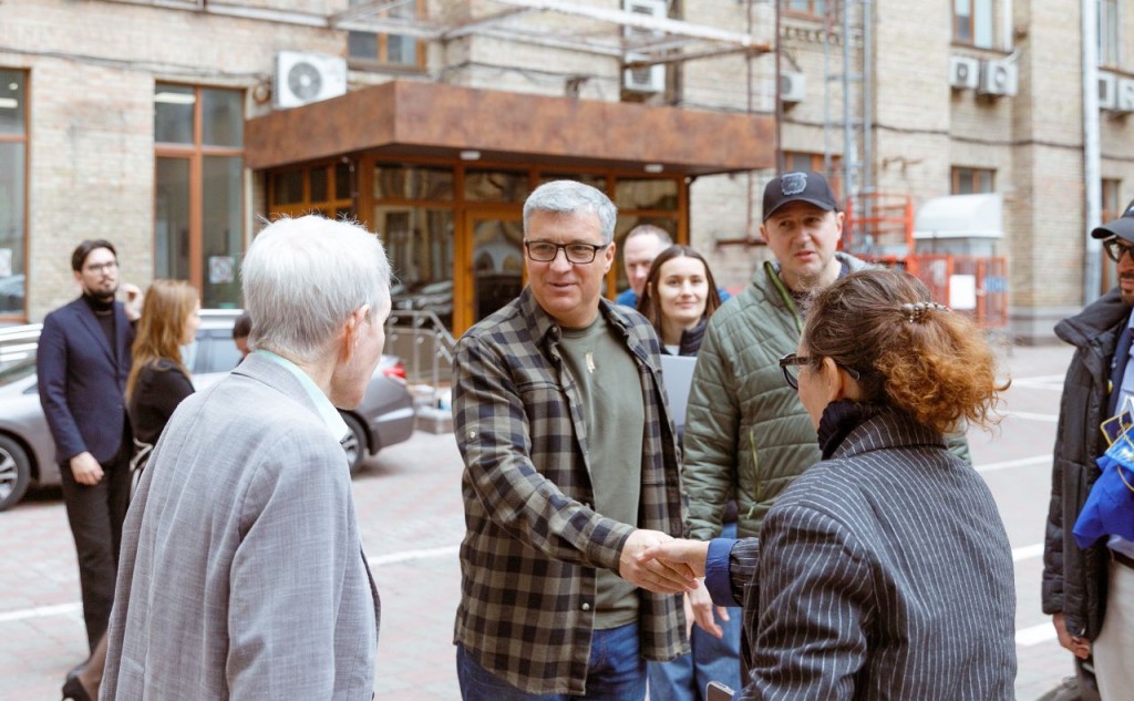 Група людей під час зустрічі на вулиці; чоловік у картатій сорочці тисне руку жінці, інші учасники спостерігають