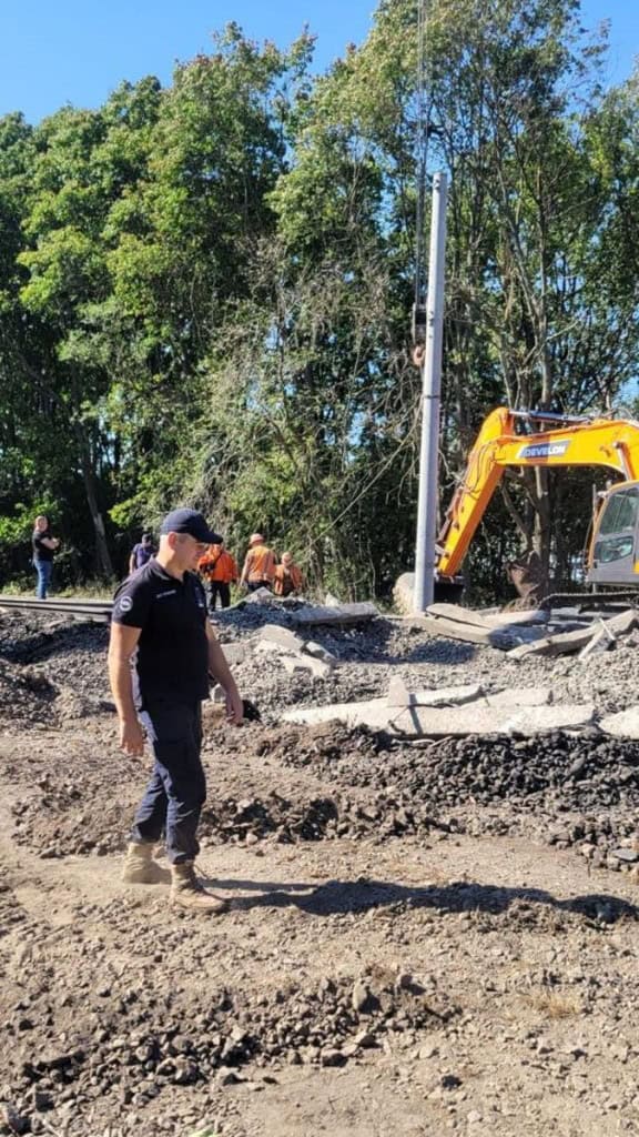 ДСНС і Укрзалізниця показали, як відновлюють рух після вибухів під Києвом (фото) Людина в чорній формі поруч із технікою, на задньому плані робітники в помаранчевих жилетах