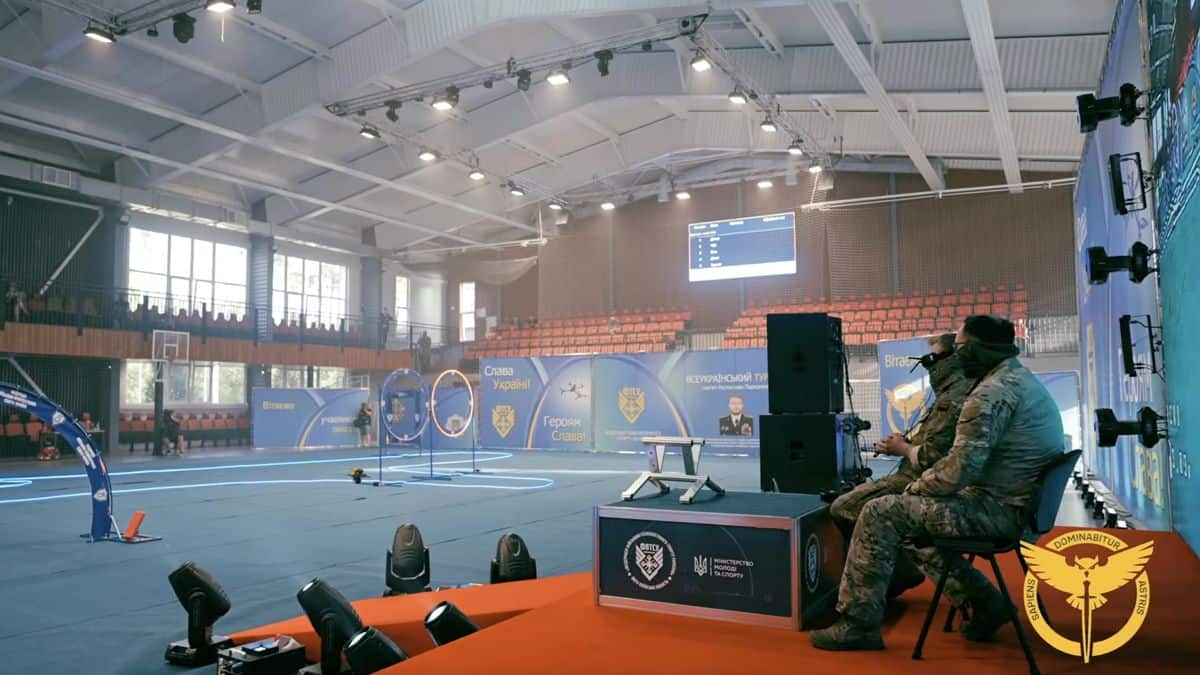 Укр: Спортивна арена в Києві під час підготовки до змагань з військових дисциплін, на передньому плані військовослужбовці в камуфляжі, сцена та фан-зона на задньому плані.