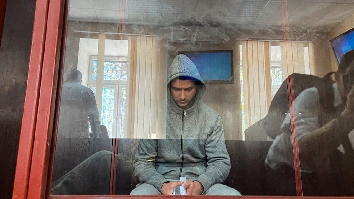 Молодий чоловік у сірому спортивному костюмі з капюшоном сидить у скляному боксі суду