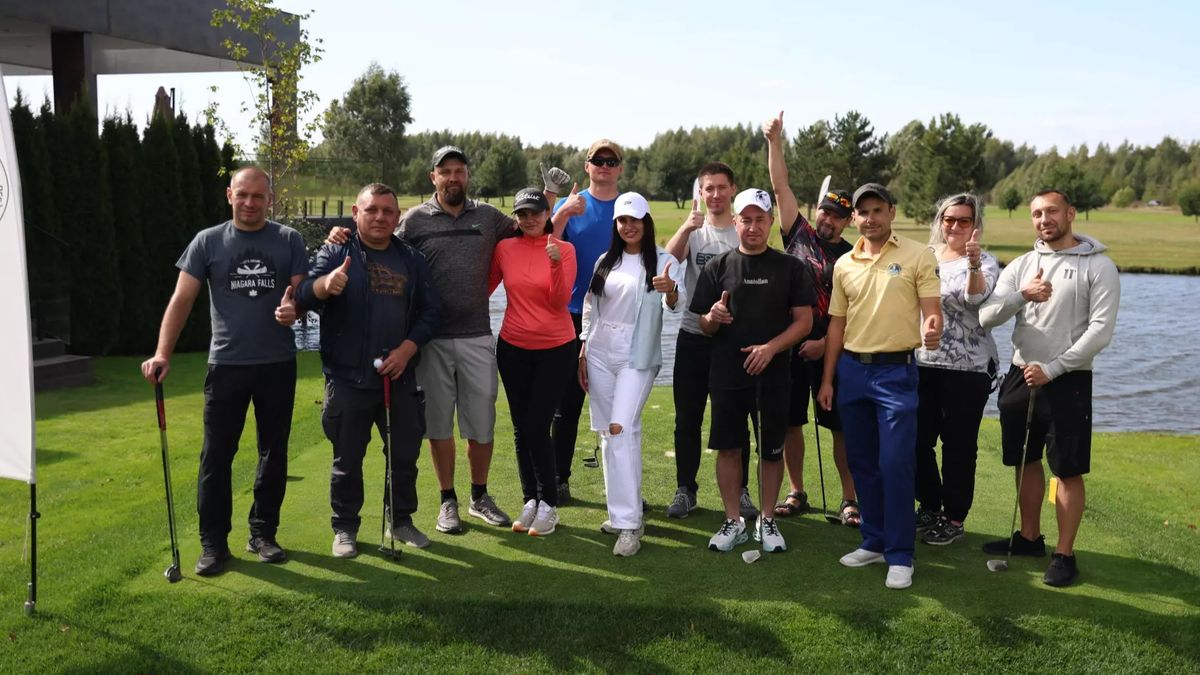 Група людей із гольфовими ключками позує для спільного фото на зеленому полі біля водойми