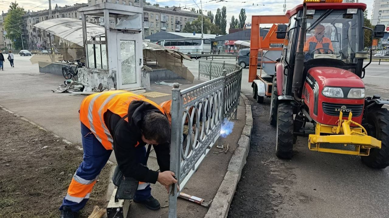 Трактор стоїть на дорозі, двоє ремонтників лагодять огорожу біля метро «Святошин» після ракетного обстрілу