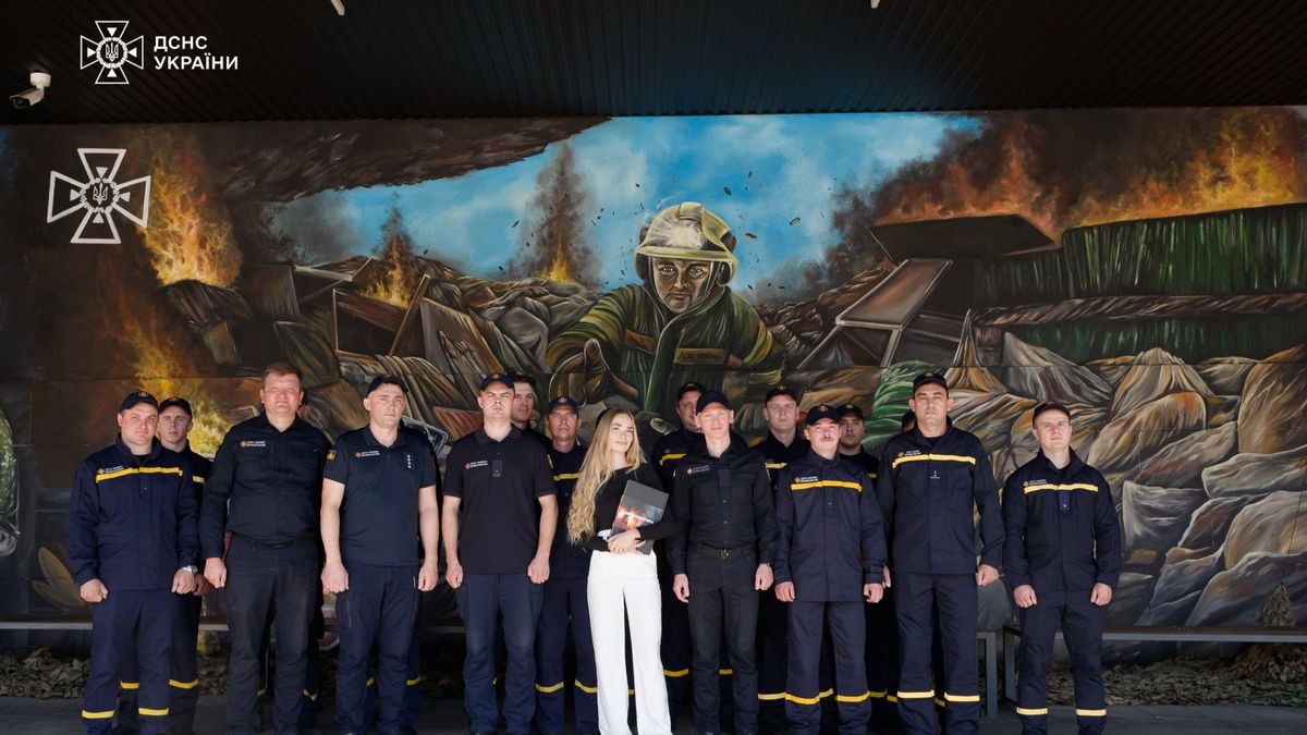 Група українських рятувальників у формі стоїть перед стіною з великим муралом, на якому зображений пожежник