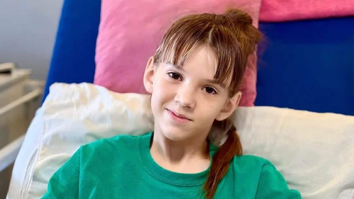 Маленька дівчинка з рудим волоссям у зеленій кофті посміхається, сидячи на ліжку з яскравою обкладинкою в кімнаті.