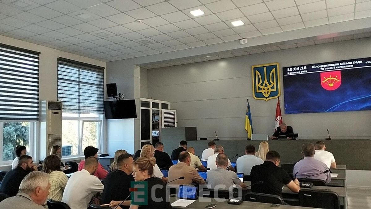 Засідання міської ради Києва в сучасній залі з українським прапором та гербом на стіні. Люди слухають доповідача, обговорюють важливі питання міста.