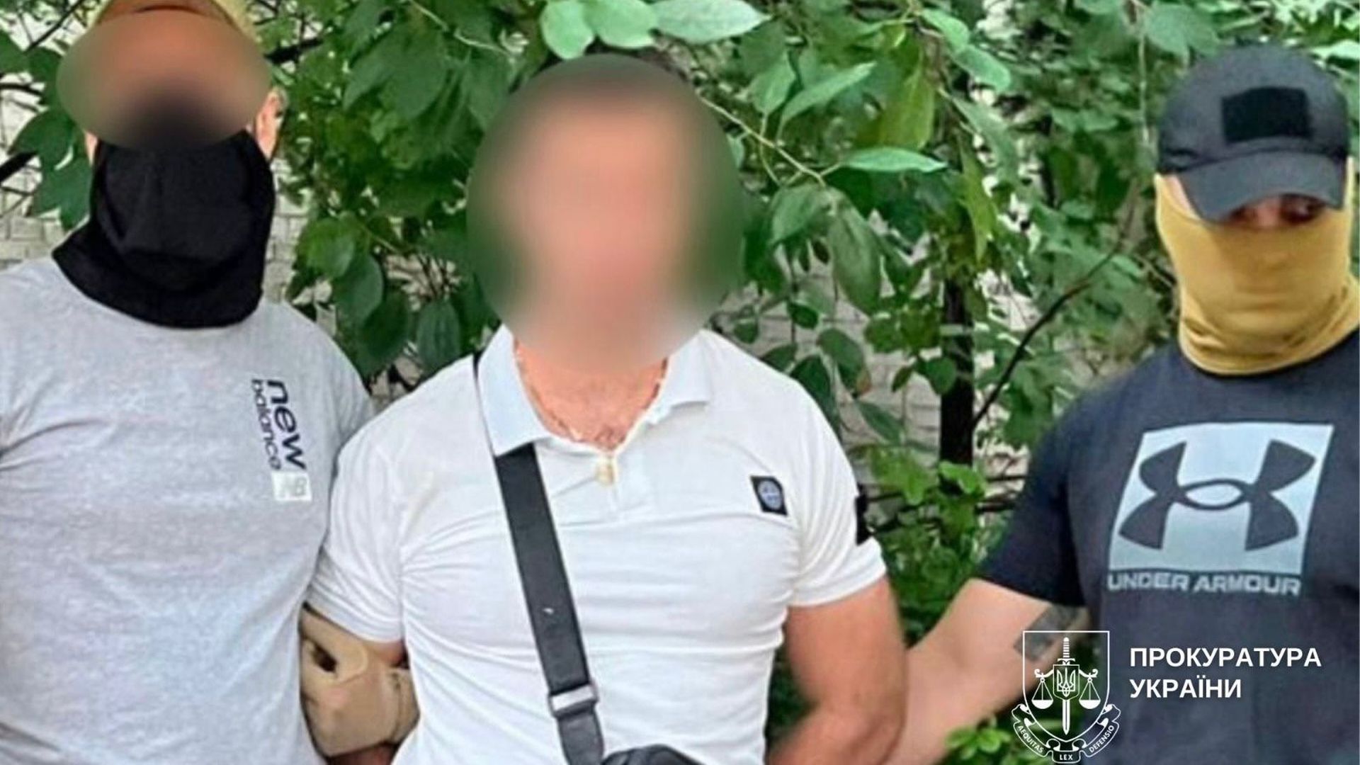 У центрі чоловік у білій футболці з розмитим обличчям, по обидва боки від нього стоять двоє правоохоронців у масках