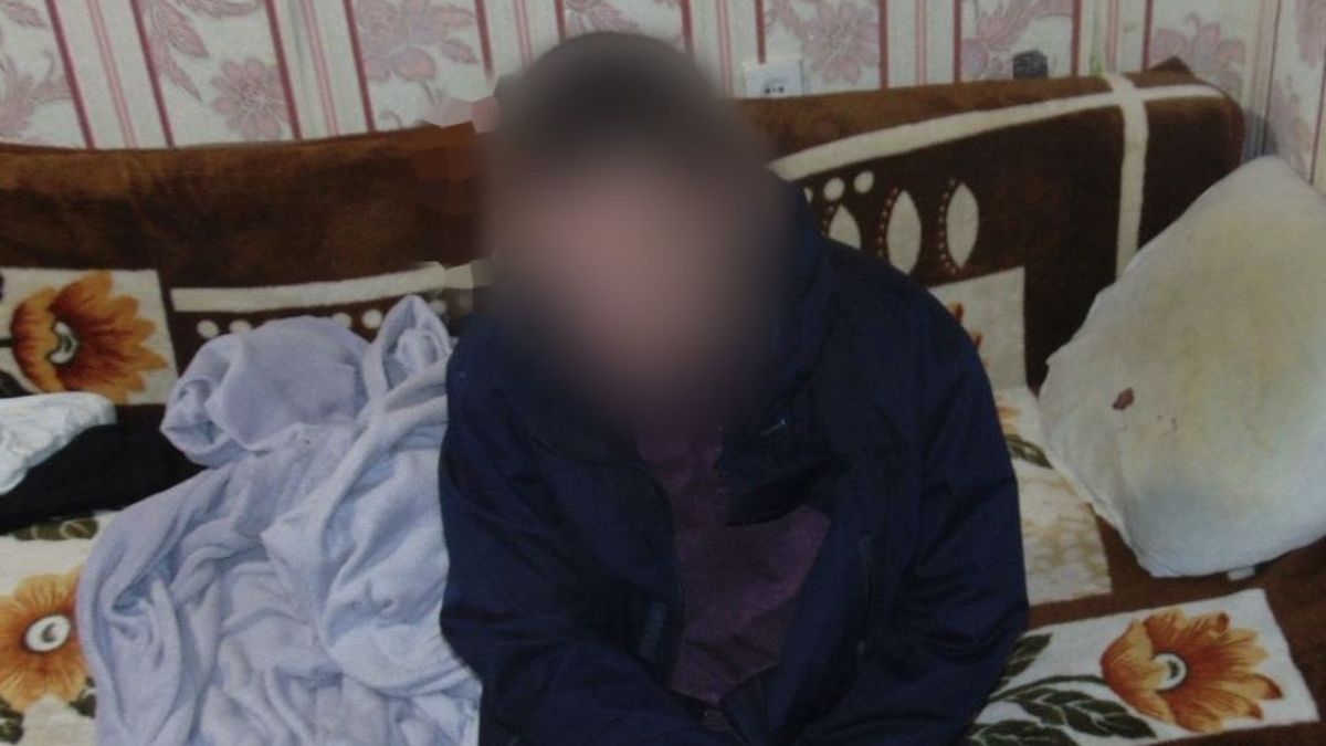 На фото чоловік у темному одязі сидить на старому дивані з квітчастим покривалом у кімнаті з блідими шпалерами