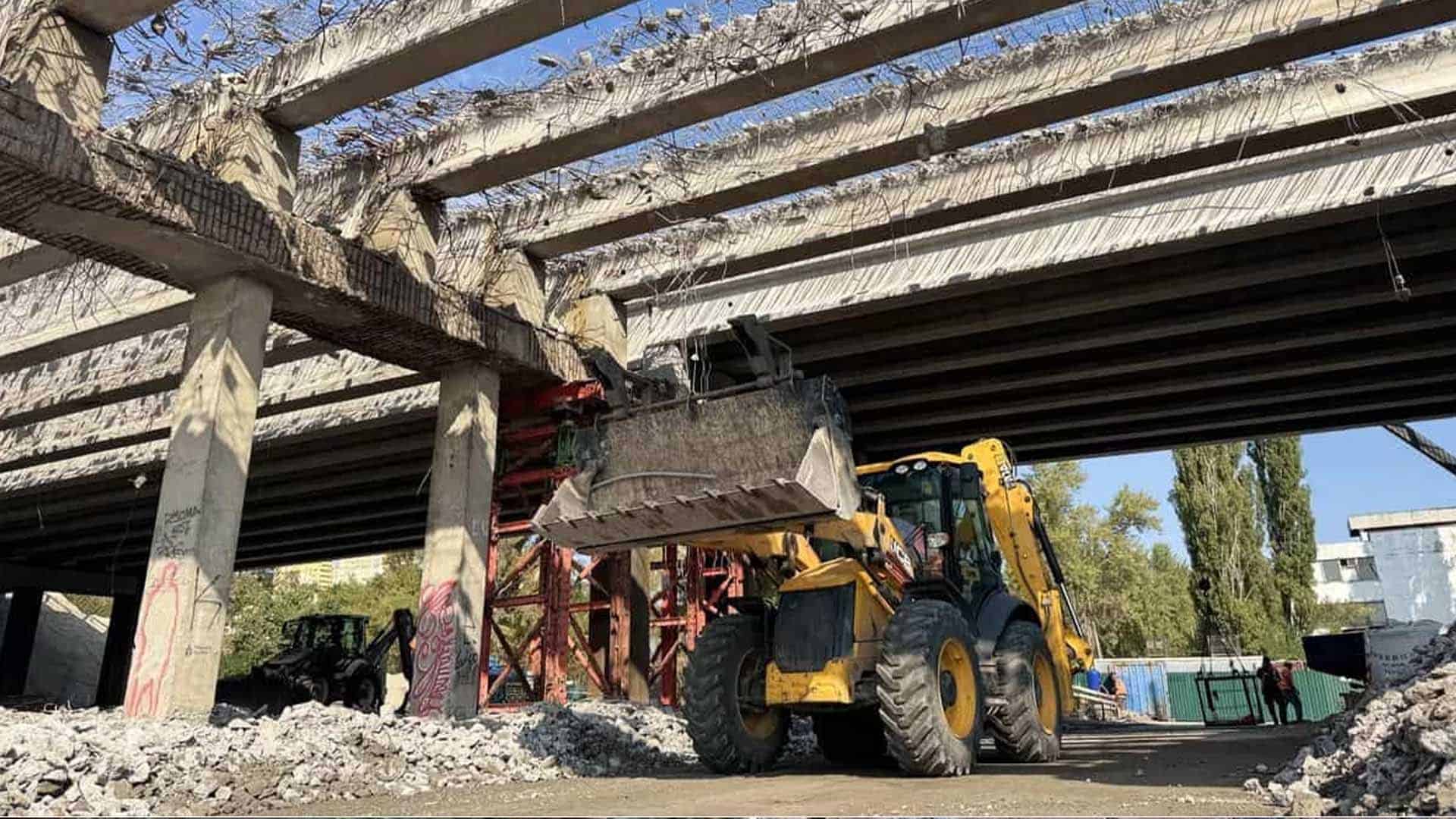 жевтий ескаватор під мостом під час демонтажу бетонних балок, з яких стирчить арматура