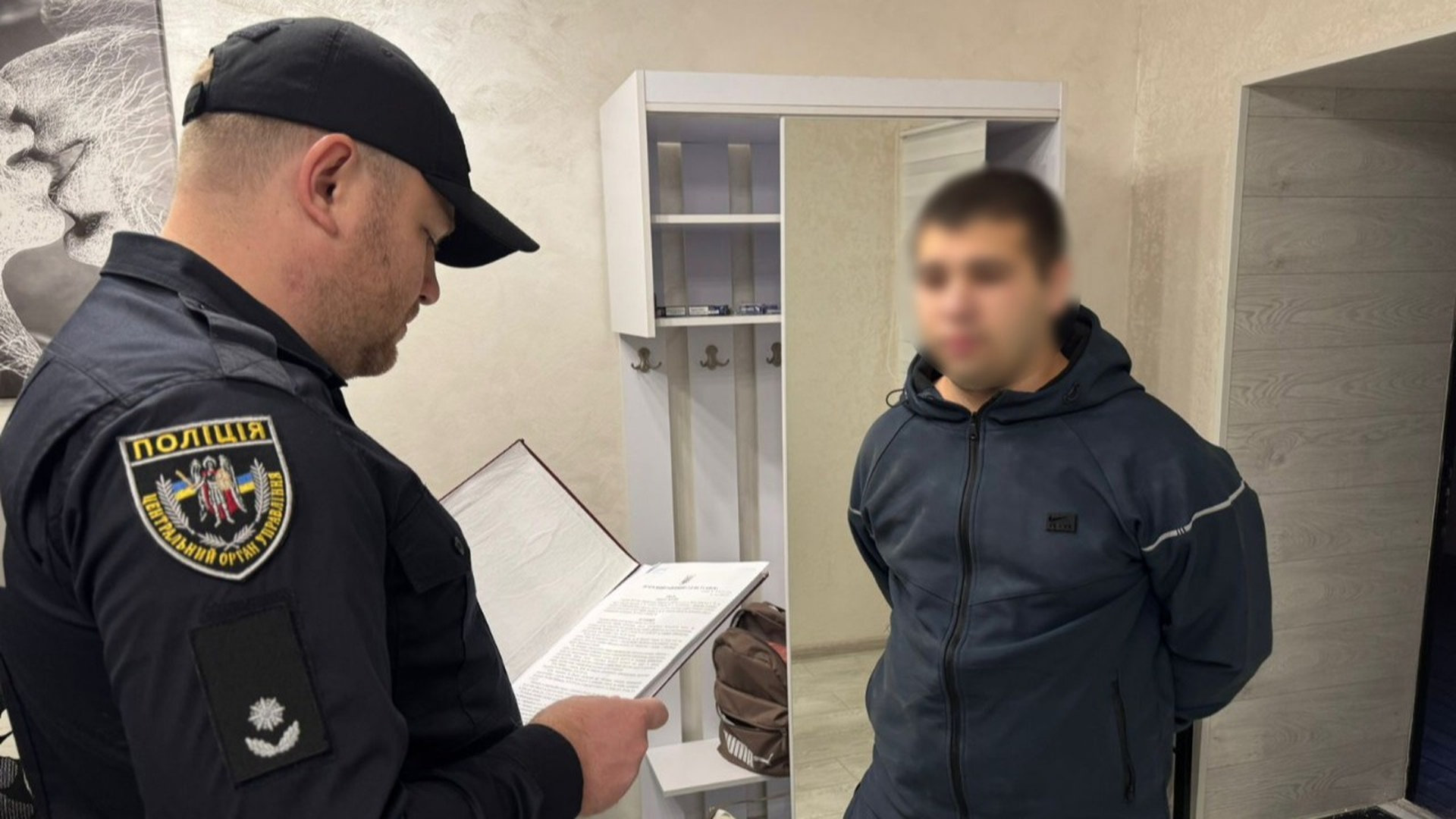 Працівник поліції стоїть з документами біля чоловіка, якого підозрюють у злочині, вони обидвоє перебувають в кімнаті будинку