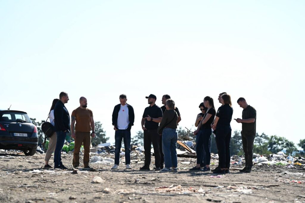 Звичайна група людей, що стоять науковоко у зоні сміття або стихійного сміттєзвалища біля автомобіля, з фоном небо та деревами, ймовірно, під час екологічної акції або огляду.