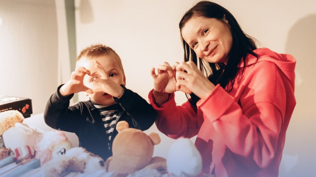 Жінка та хлопчик складають руками фігурку серця, сидячи серед м’яких іграшок