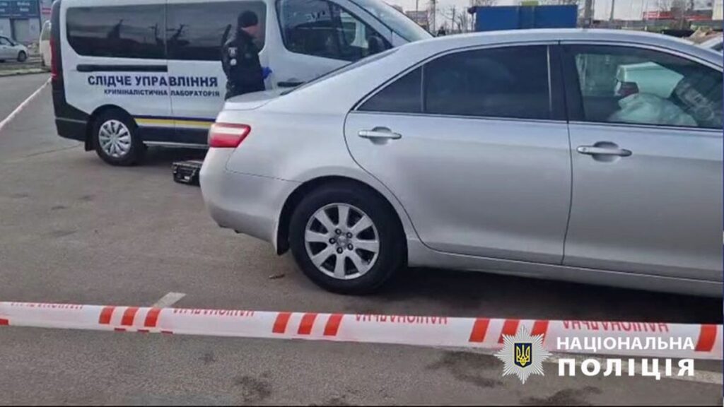 Сірий легковий автомобіль та мікроавтобус криміналістичної лабораторії поліції, огороджені стрічкою на парковці