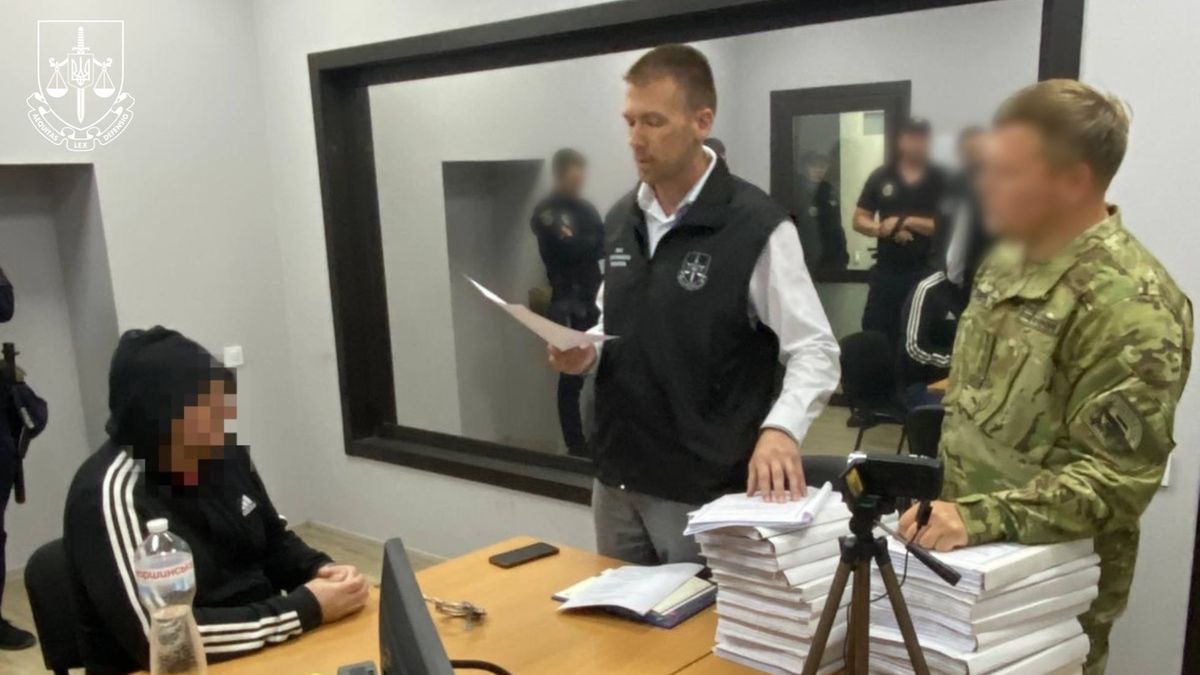 Чоловік у чорній кофті з капюшоном сидить за столом, поруч прокурор зачитує документи