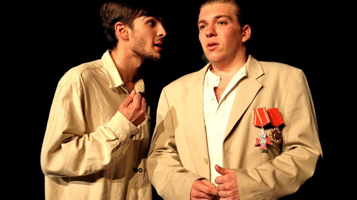 Двоє акторів на театральній сцені: один у світлій сорочці говорить, нахилившись до іншого