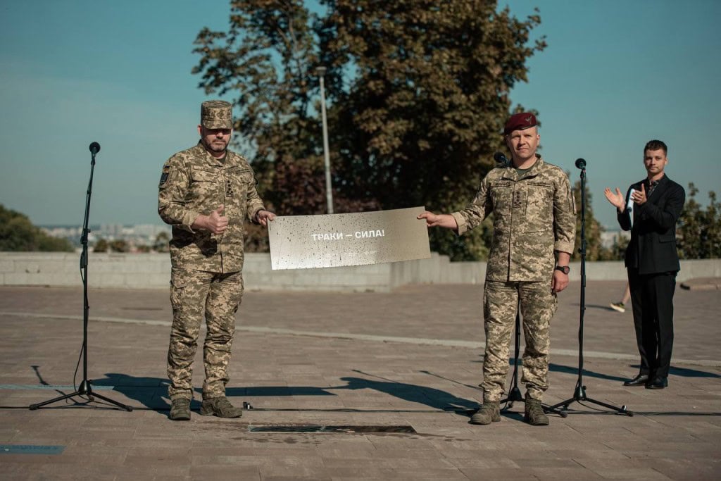 На Алеї захисників у Києві відкрили пам’ятний знак воїнам-танкістам Двоє українських військових у камуфляжі тримають металеву табличку з написом «ТРАКИ — СИЛА!»