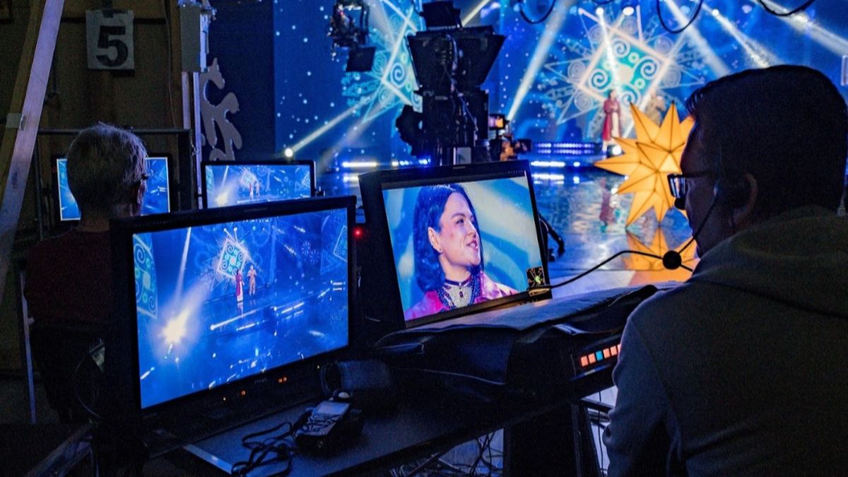 Яскраво освітлена телевізійна студія: на передньому плані пульти та монітори, де відображаються крупний план ведучої та сцена