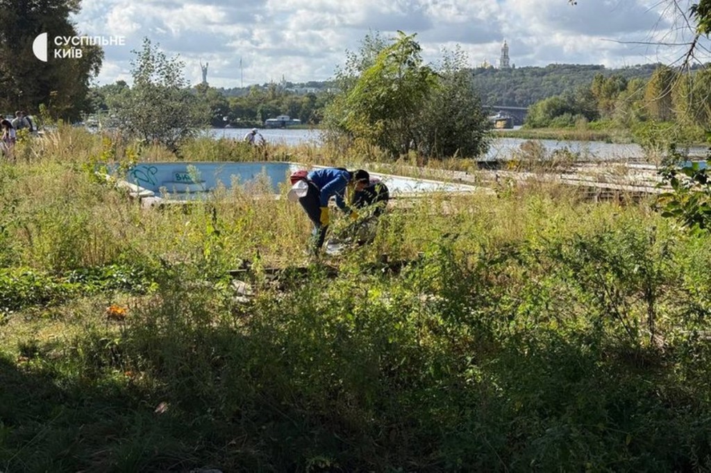 Волонтери збирають сміття серед заростей біля річки, на фоні видно Києво-Печерську лавру